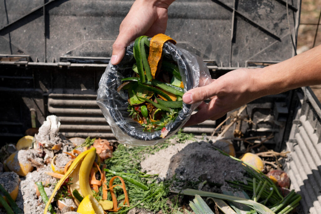 Una persona añade restos de comida, como cáscaras de plátano y verduras, a un recipiente de reciclaje al aire libre lleno de materia orgánica en descomposición.