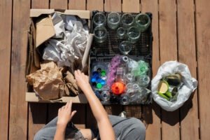 Una persona clasifica materiales reciclables: cartón y papel en una caja, vidrio en otra, y plásticos y latas en bolsas separadas.