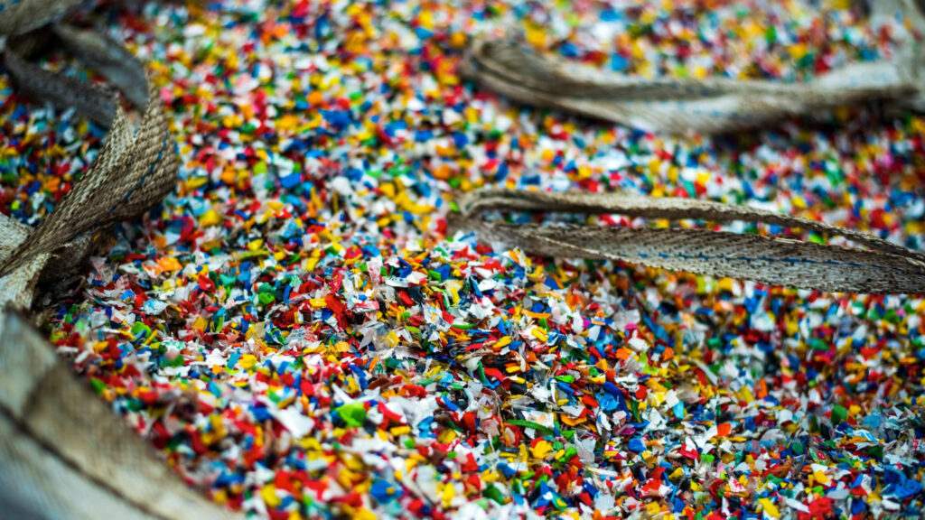 Una bolsa de yute abierta llena de gránulos de plástico reciclado de varios colores, como parte de un proceso de reciclaje mecánico.