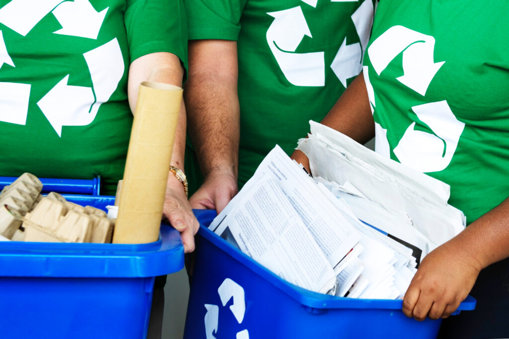 Vista de las manos de personas con playeras verdes que colocan papeles y cartón, dentro de contenedores de reciclaje. 