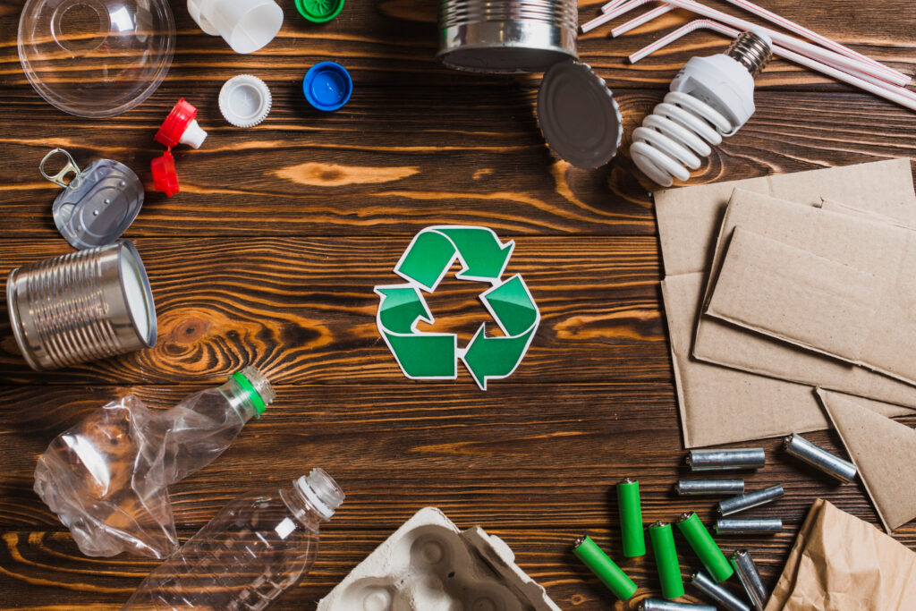 Una vista aérea de varios materiales reciclables, como plástico, metal, papel y baterías, rodeando un gran símbolo de reciclaje verde sobre una mesa de madera.